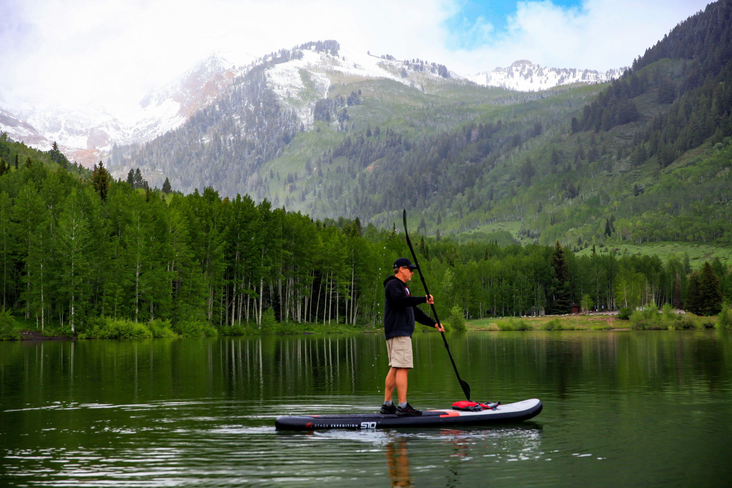 Matt paddleboard silver lake res A person paddleboards on a calm lake surrounded by lush green trees and mountains with patches of snow under a partly cloudy sky.