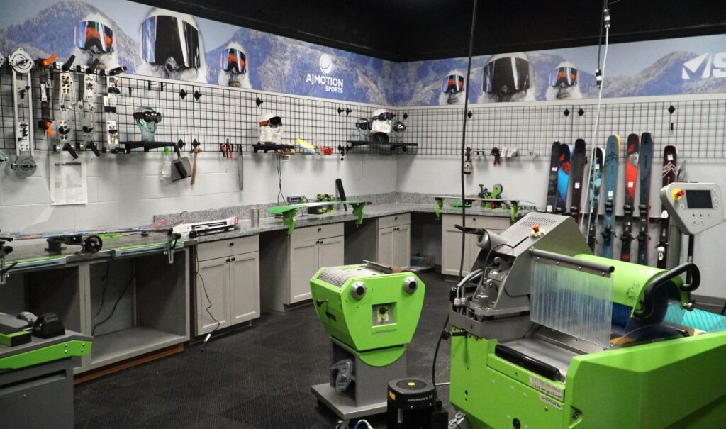 A ski and snowboard tuning workshop with green tuning machines, workbenches, wall-mounted tools, and several pairs of skis and snowboards lined up along the wall. Mountain-themed posters decorate the upper walls.