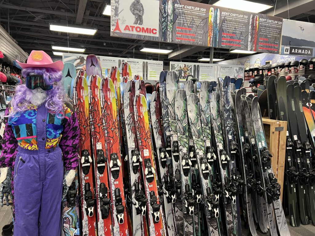 Mannequin in a pink cowboy hat, purple wig, and colorful outfit stands next to racks of colorful skis inside a ski shop, with brand signs like Atomic and Armada visible overhead.