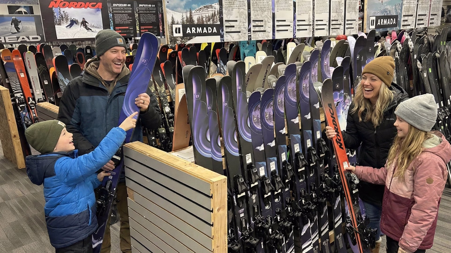 armada_family_shopping_1920x1080 A smiling family of four, dressed in winter clothing, looks at skis together inside a ski shop. The parents and two kids are surrounded by rows of upright skis and ski gear displays.