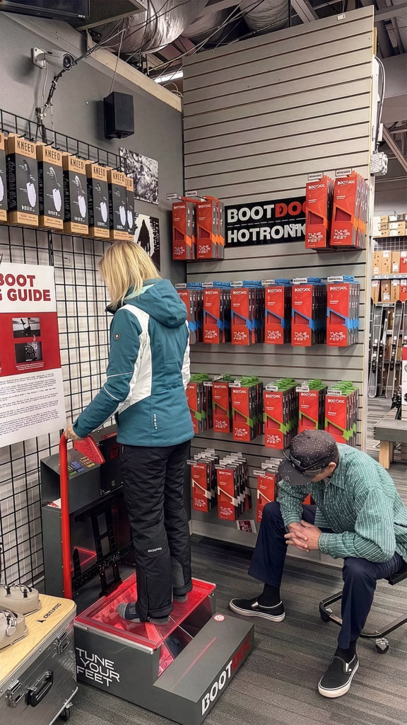 bootdoc_footbed_gemini_1080x1920 A woman in a teal jacket stands on a ski boot fitting machine, while a man crouches nearby checking her boots. Behind them are shelves filled with ski boot insoles and accessories.