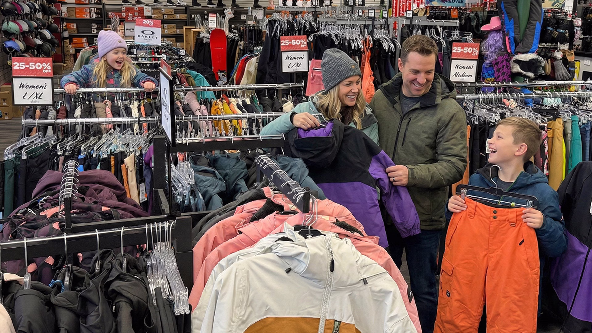 clothing_union-square_family-shopping_1920x1080 A family of four wearing winter clothes smiles and shops for ski outfits in a store; racks of colorful jackets and snow pants surround them, with sale signs visible above the racks.