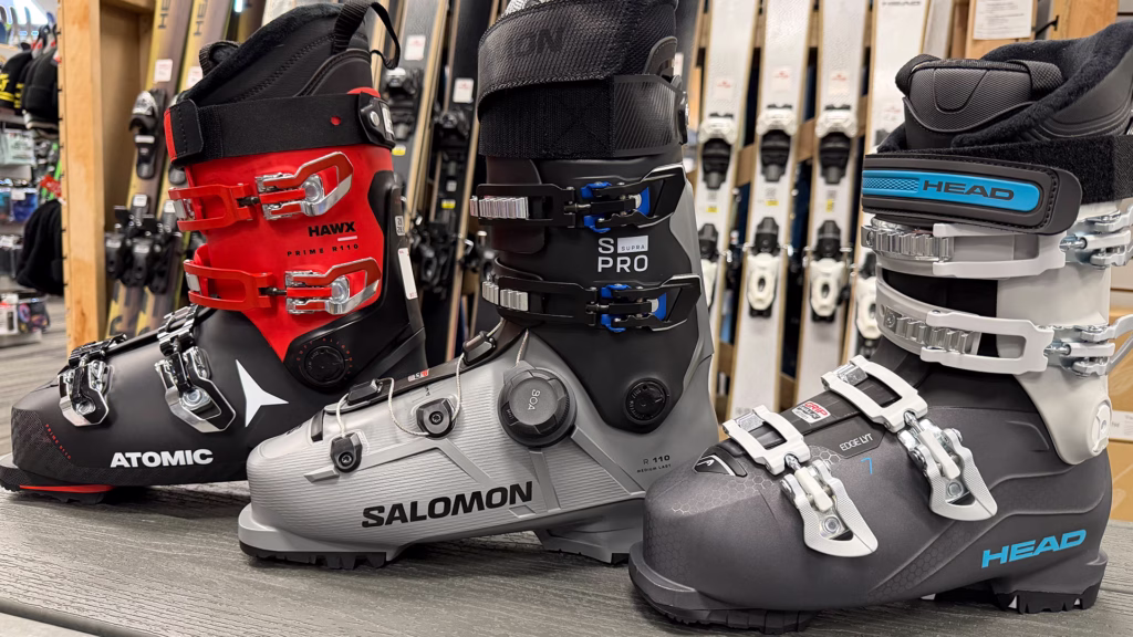 featured_ski_boots_1920x1080 Three ski boots—Atomic (red and black), Salomon (gray and black), and Head (gray and blue)—are displayed side by side on a bench in a ski shop, with ski equipment in the background.