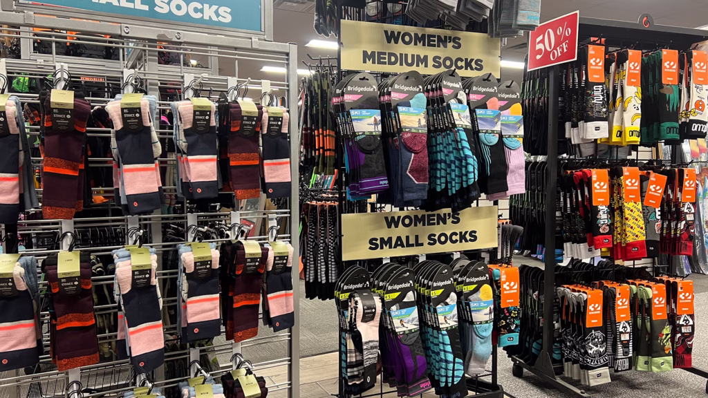 ski_sock_racks_in-store_aspen_plaza_1920x1080 A store display with racks of colorful women’s socks labeled “Women’s Small Socks” and “Women’s Medium Socks.” A “50% Off” sale sign hangs above more socks on the right, with various brands and patterns visible.