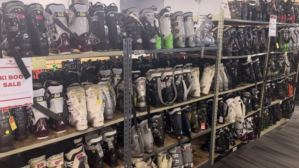 used_ski-boots_union-square_1920x1080 Shelves filled with various pairs of ski boots are organized by size and style in a store. A sale sign is visible on the left, and the floor has a gray patterned carpet.