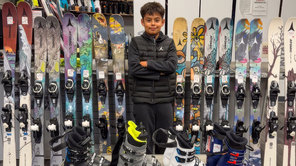 A teenage boy skier standing in front of a line of jr demo skis from Atomic